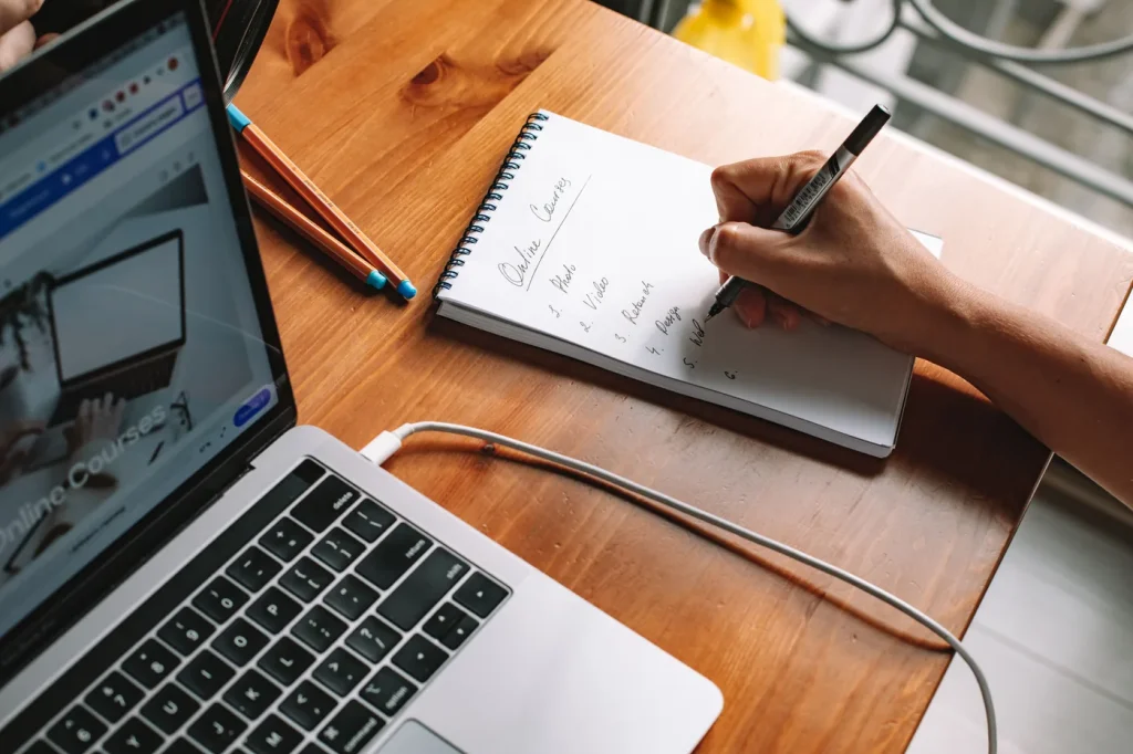 A person making notes beside a laptop during online research.
